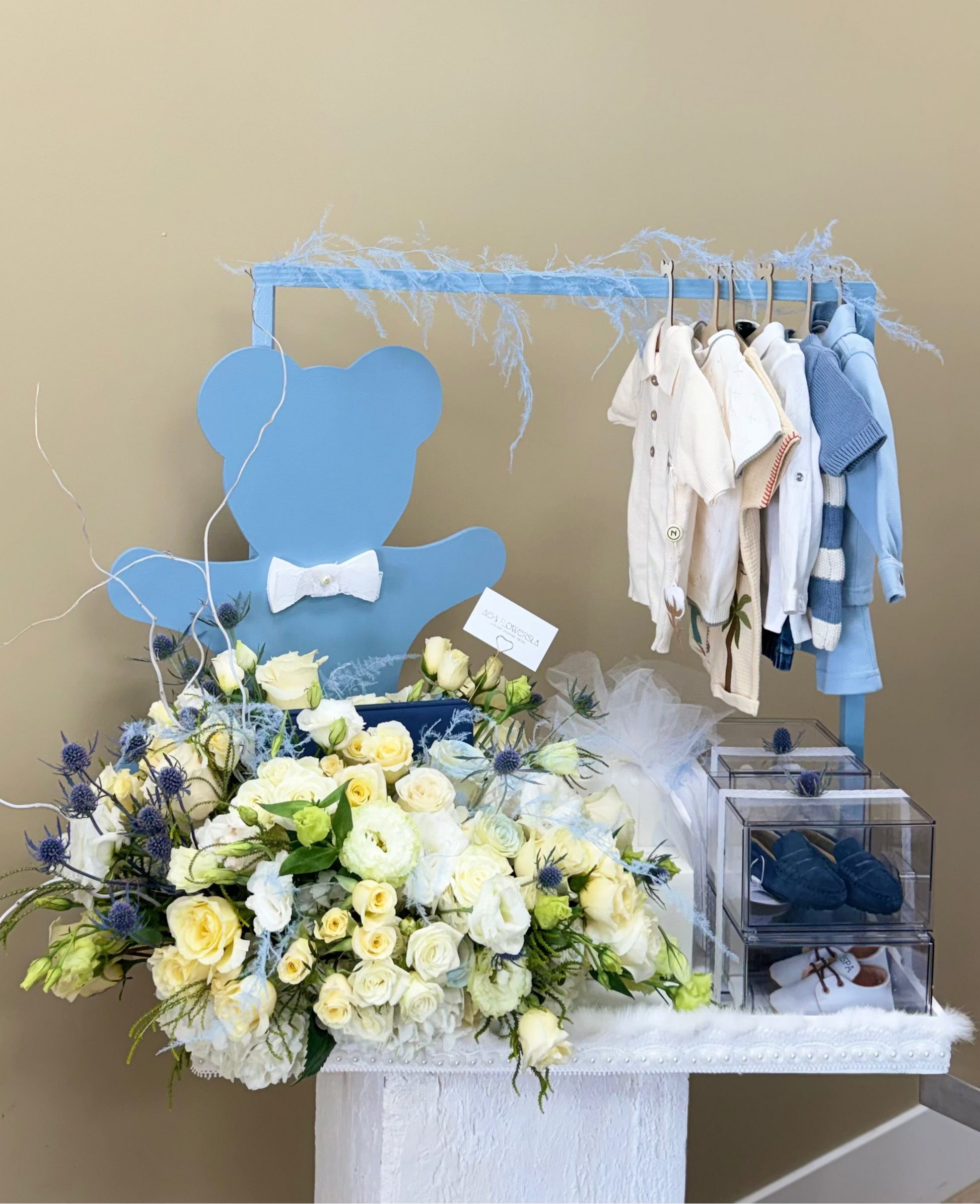 A baby shower or first birthday celebration display featuring a light blue wooden bear silhouette with a white bow tie, a clothing rack with white and blue baby outfits, a large bouquet of white and yellow roses, and a clear box containing blue shoes.
